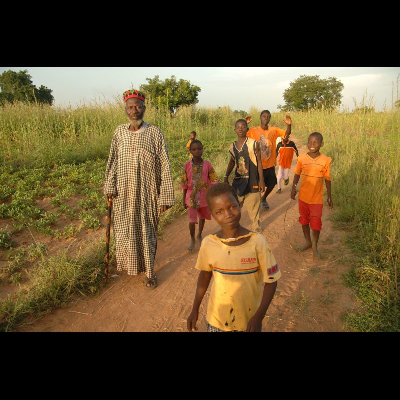 SABTENGA, Burkina Faso — Musanai Zemnai, the Chief of the Young People, walks through the bush village of Sabtenga, with children surrounding him. A small outreach group has recently been started in the town, and Zemnai welcomed the visitors as part of the local leadership.