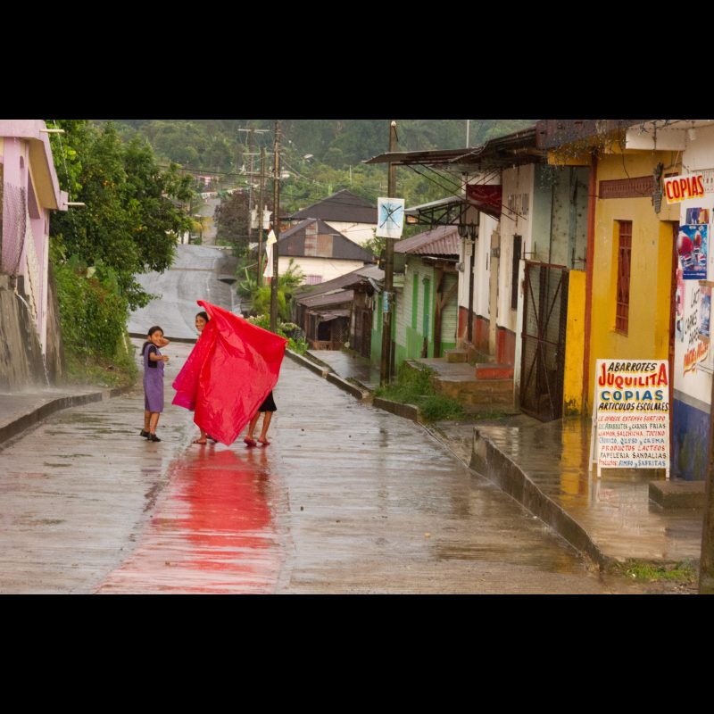 SALVADOR URBINA, Mexico — A mother and her two daughters glance back as they use a tarp to shield themselves from the rain while running home in Salvador Urbina, Chiapas, Mexico. Sudden downpours are common in this mountainous region, where families often rely on quick improvisation.