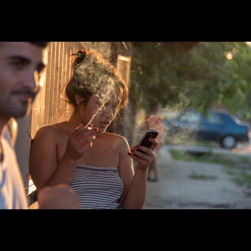 HERĂȘTI, Romania — A young woman smokes a cigarette while checking her phone on a quiet street in Herăști, Giurgiu County, Romania. Daily life in rural villages blends traditional lifestyles with modern habits.