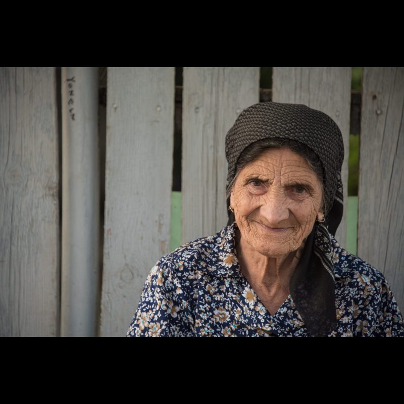 An elderly woman smiles warmly outside her home in Herăști, Romania. Her traditional headscarf and patterned blouse reflect the rural customs of the region, while her expression offers a quiet glimpse into a life shaped by resilience and community.